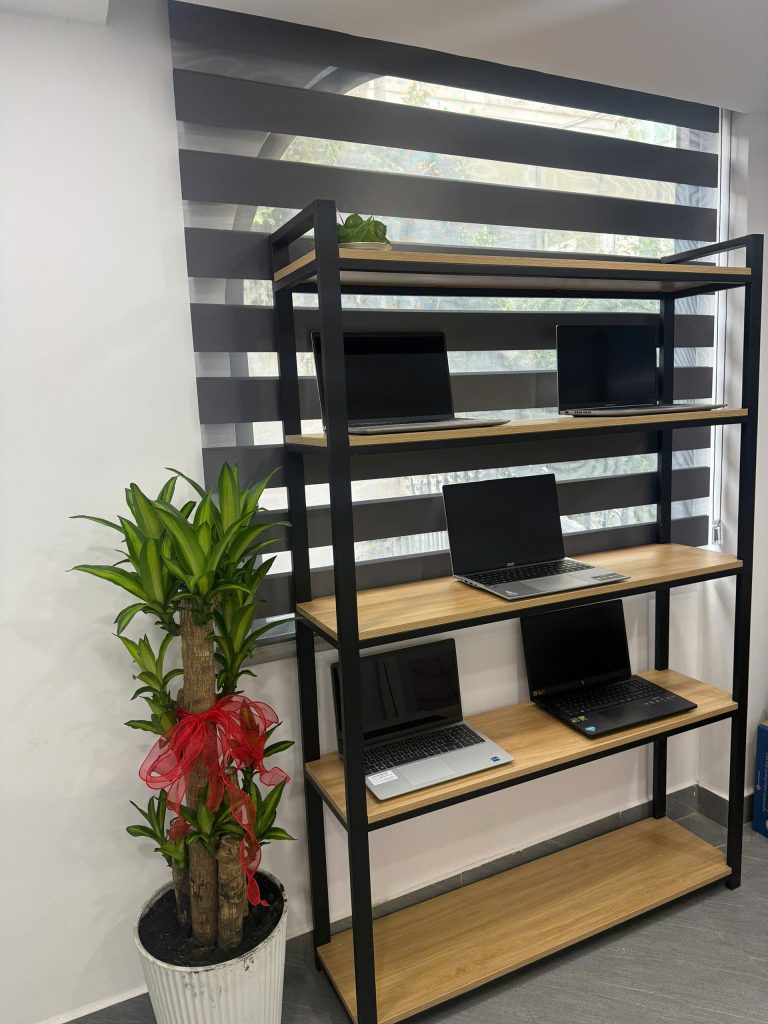 Kệ khung sắt đen mặt gỗ ở chi nhánh 2 trưng bày năm chiếc laptop mở nắp, bên cạnh có chậu cây cảnh
